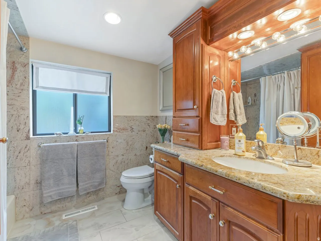 Recessed lighting, wood-finish cabinetry, stone countertop, tile flooring, and a large mirror with integrated lighting