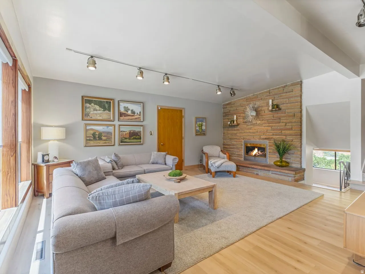 Stone-clad fireplace with hearth, track lighting, wood-finish flooring, and a large window
