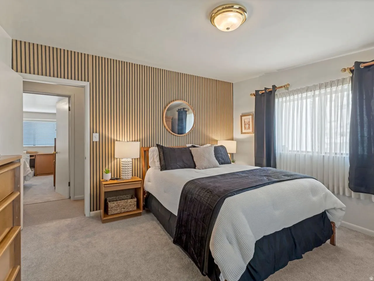 Textured accent wall, neutral carpeting, window with privacy blinds, flush mount ceiling light, and built-in bedside table with lamp