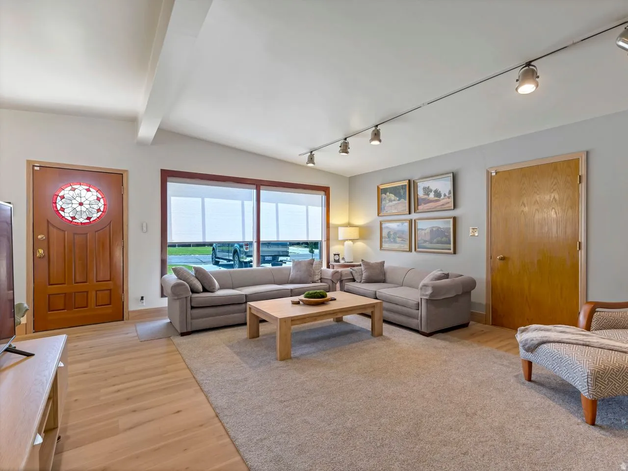 Vaulted ceiling with exposed beams, wood-finish flooring, track lighting, and a large picture window
