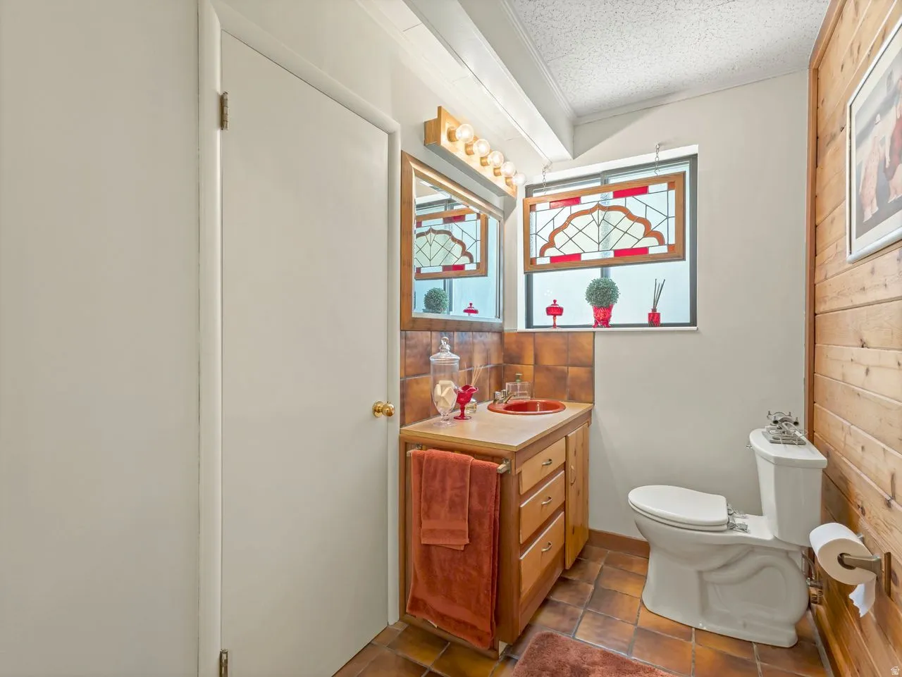 Window with decorative stained glass, wood-panel accent wall, wood-finish vanity with countertop, tiled flooring, and a white toilet