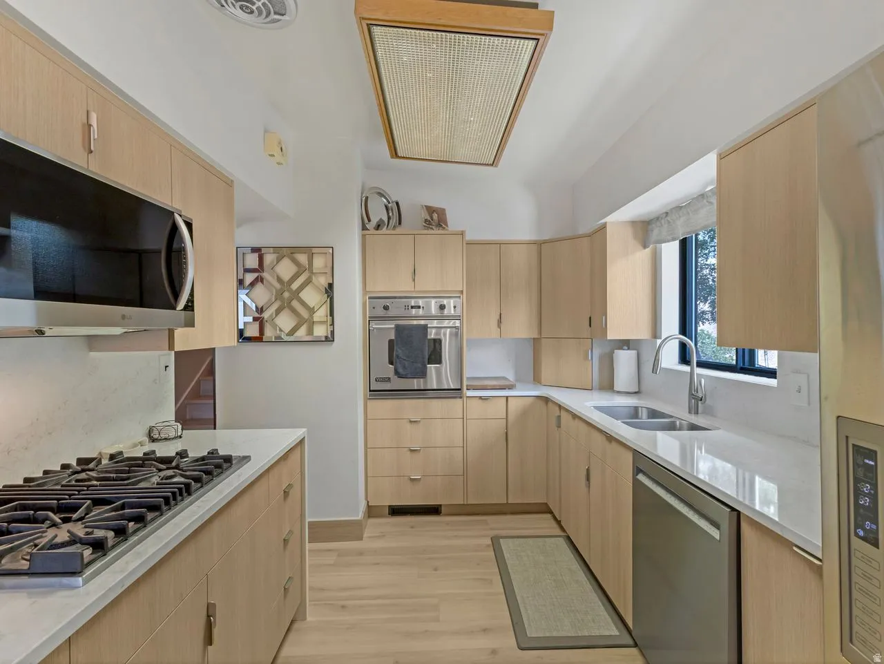 Light wood-finish cabinetry with modern hardware, stainless steel appliances, light-toned countertops, wood-finish flooring, and a rectangular window