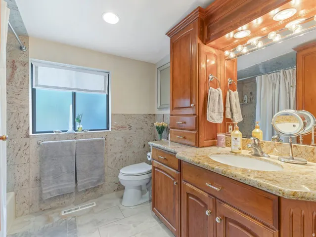 Recessed lighting, wood-finish cabinetry, stone countertop, tile flooring, and a large mirror with integrated lighting