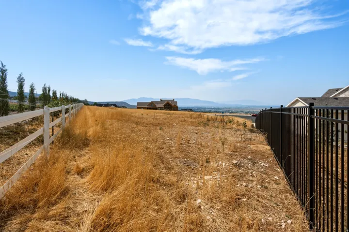 Expansive undeveloped lot featuring a white multi-rail fence and a black metal fence, with surrounding residential structures and distant mountains