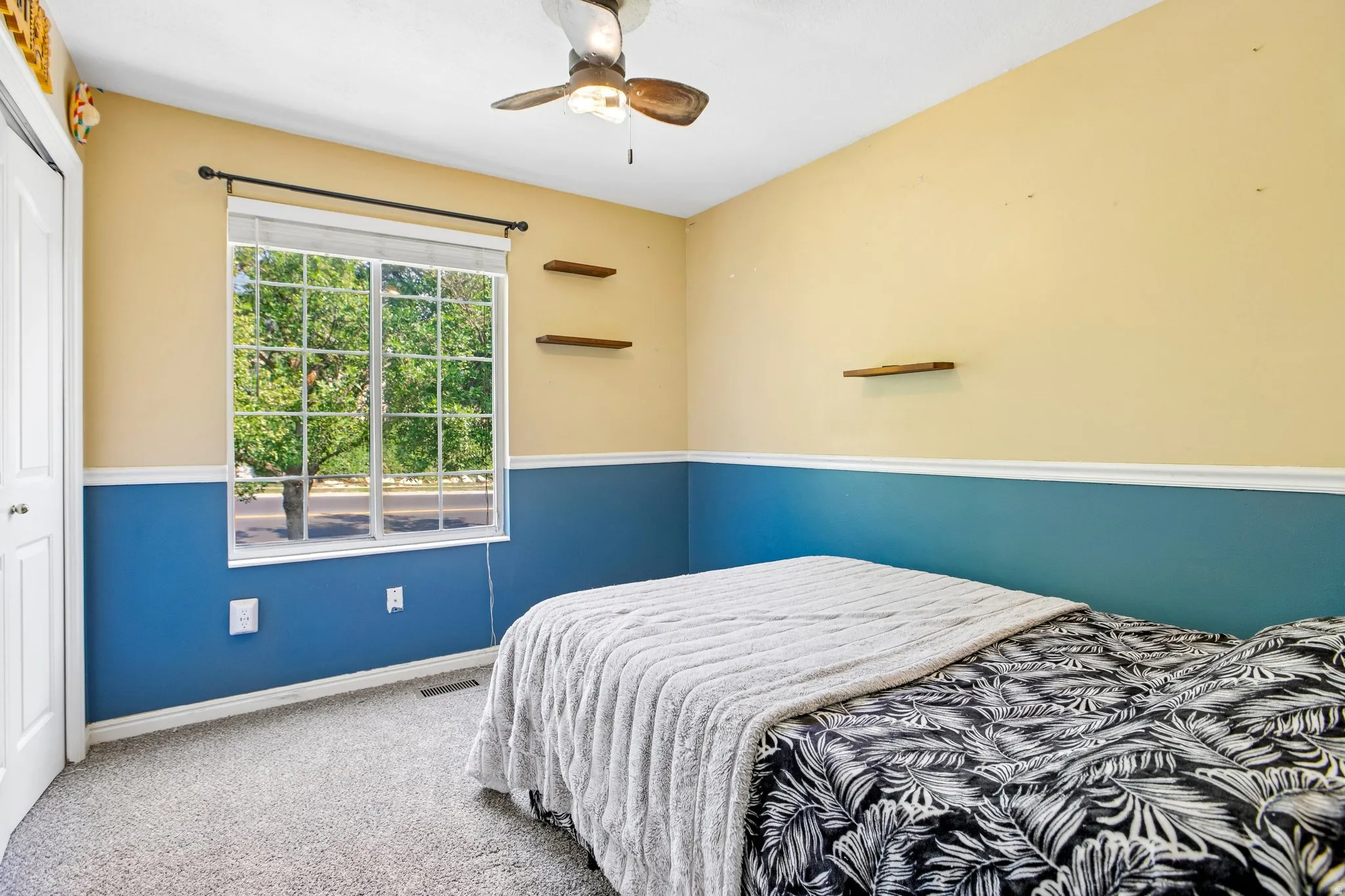 Window with horizontal blinds, overhead ceiling fan with light, wall-mounted shelving, and carpeted flooring