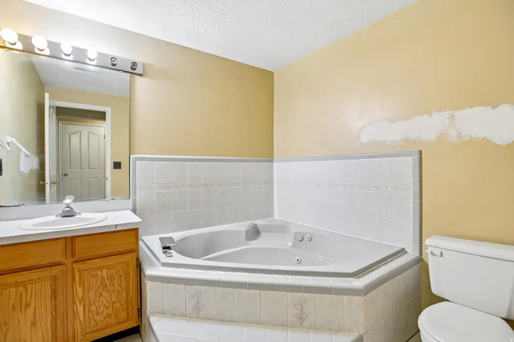 Corner soaking tub with tiled surround, wood-finish vanity with countertop sink, and a mirrored medicine cabinet with vanity lighting