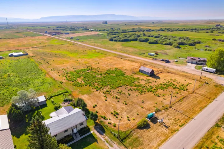 Expansive property featuring a detached house with a metal roof, a distinct outbuilding, and a gravel driveway, surrounded by open fields and distant mountains