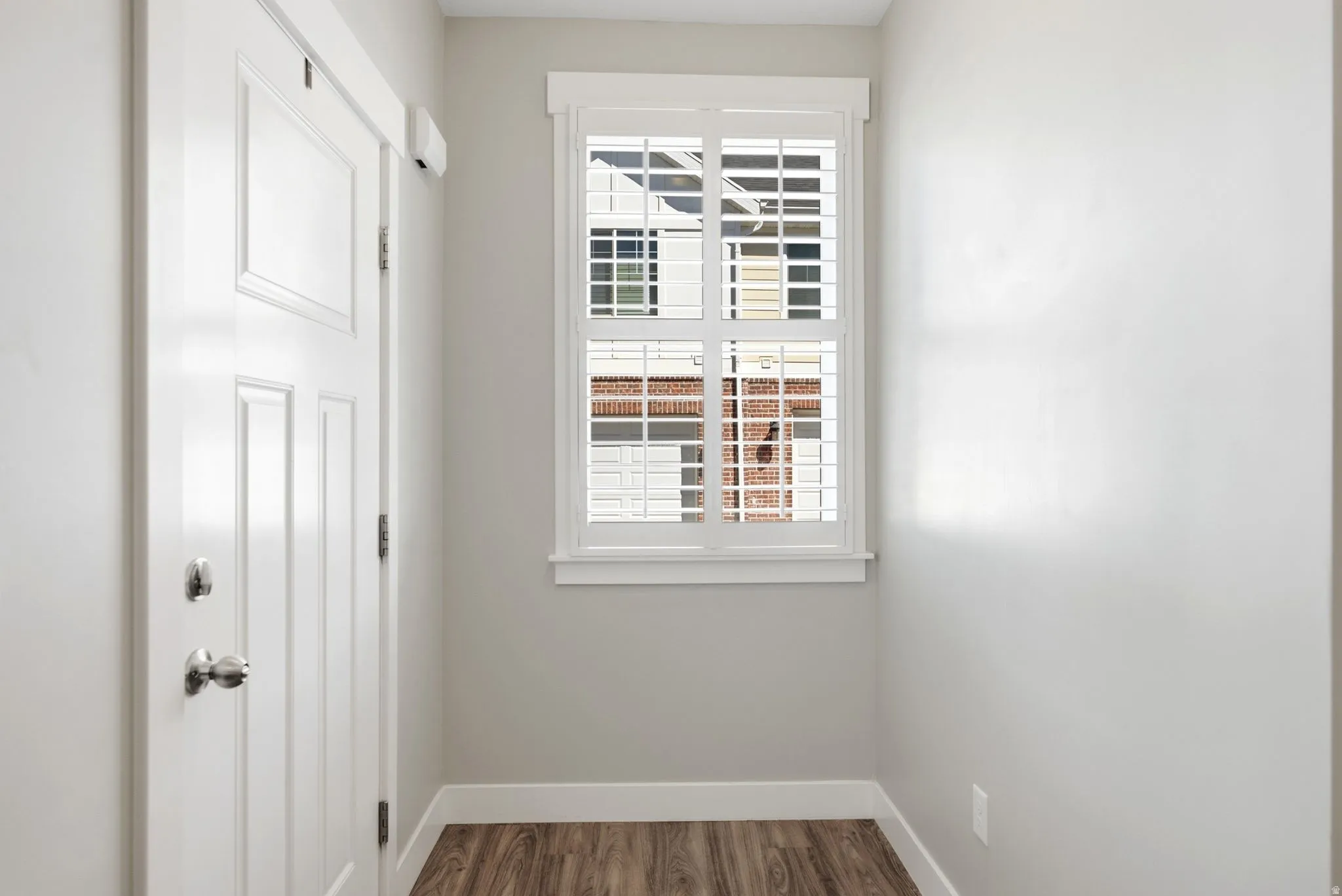 Wood-finish flooring, white paneled door with polished hardware, and a window featuring white plantation shutters