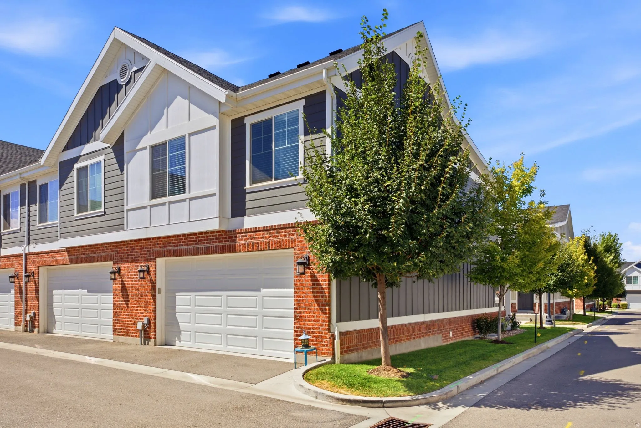 Contemporary townhome featuring a brick and siding exterior with gabled roofline, dual two-car garage doors, large multi-pane windows, and mature landscaping with established trees along the front façade