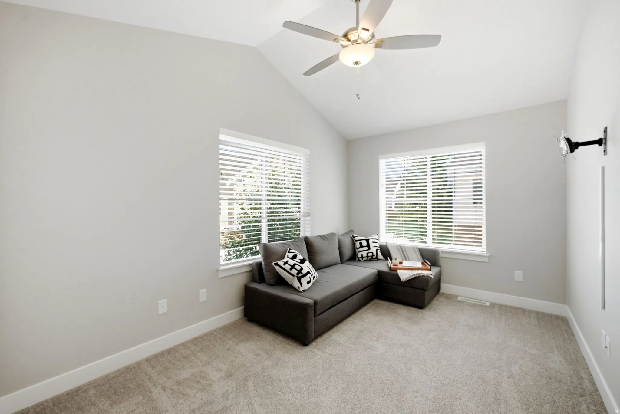 Vaulted ceiling, ceiling fan with integrated lighting, two windows with blinds, light-toned carpeting, and a wall-mounted light fixture