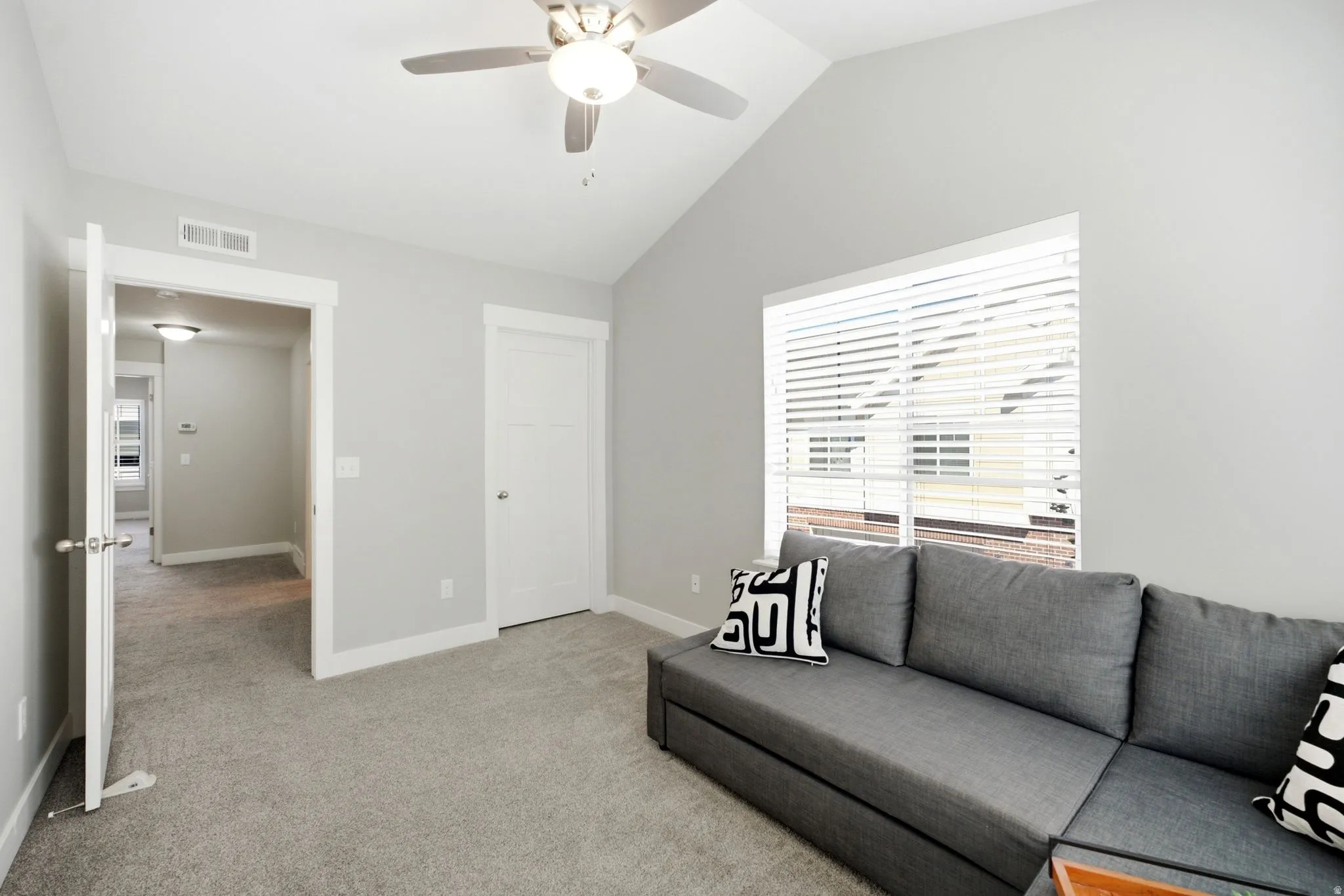 Bright secondary bedroom with vaulted ceiling, ceiling fan with integrated lighting, neutral carpet flooring, and large window with plantation shutters providing natural light