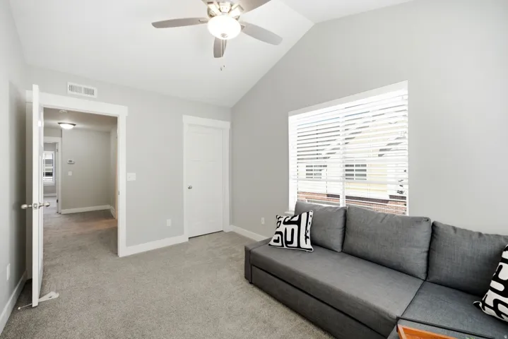 Bright secondary bedroom with vaulted ceiling, ceiling fan with integrated lighting, neutral carpet flooring, and large window with plantation shutters providing natural light