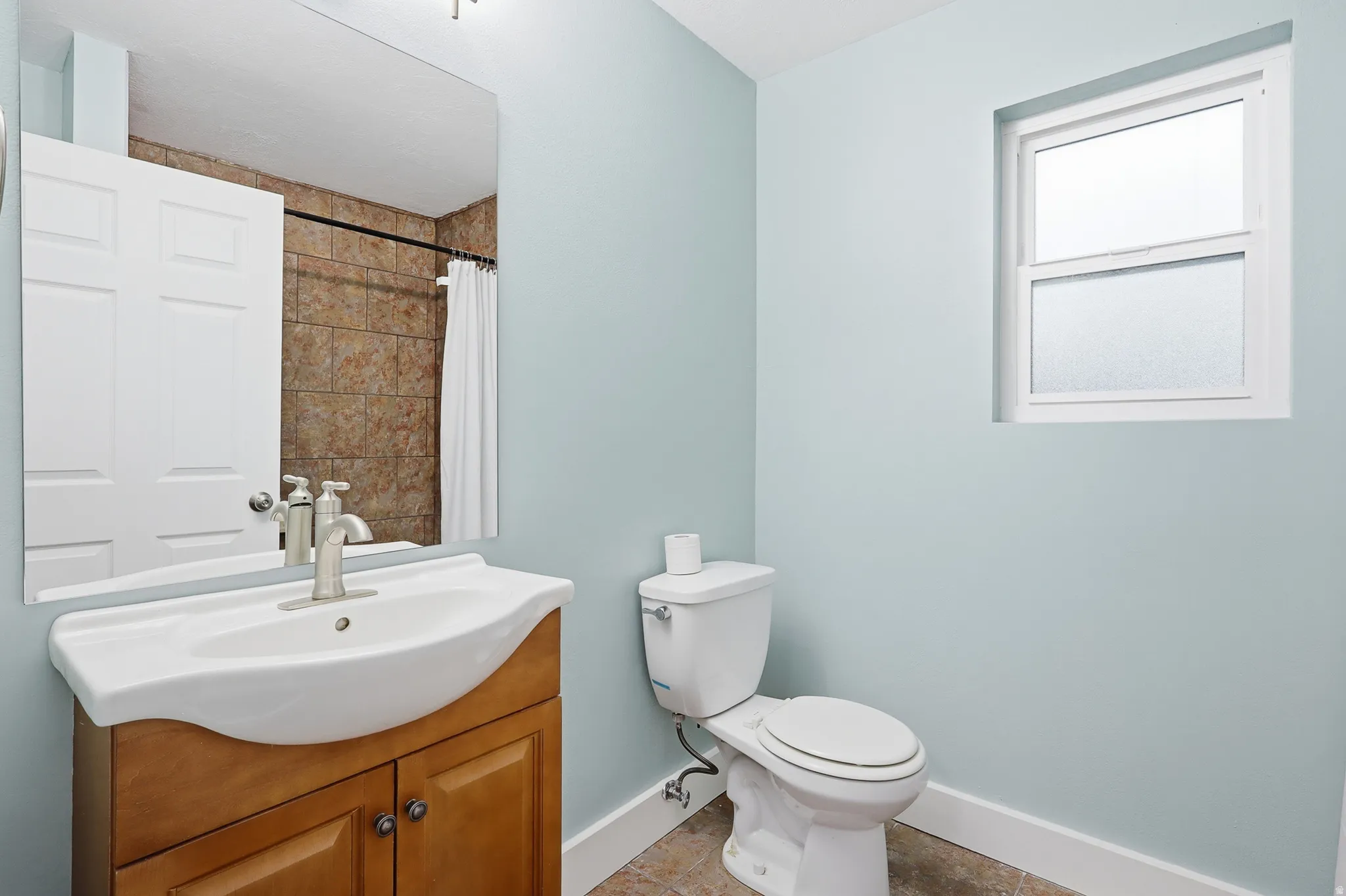 Wood-finish vanity with an integrated white sink, brushed nickel faucet, large wall mirror, and a fixed pane window