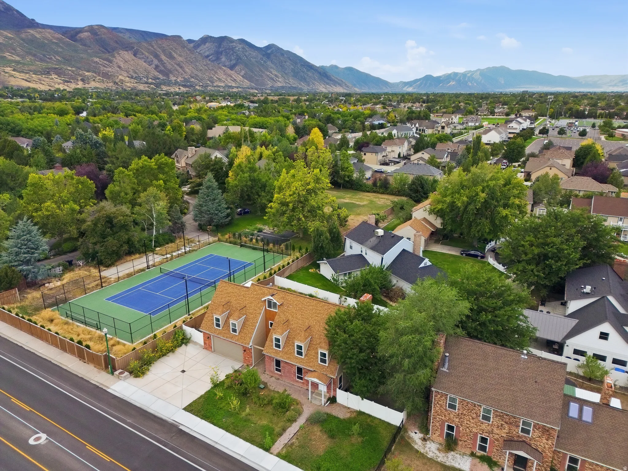 Brick exterior with dormer windows, attached garage, concrete driveway, and adjacent tennis court with fencing