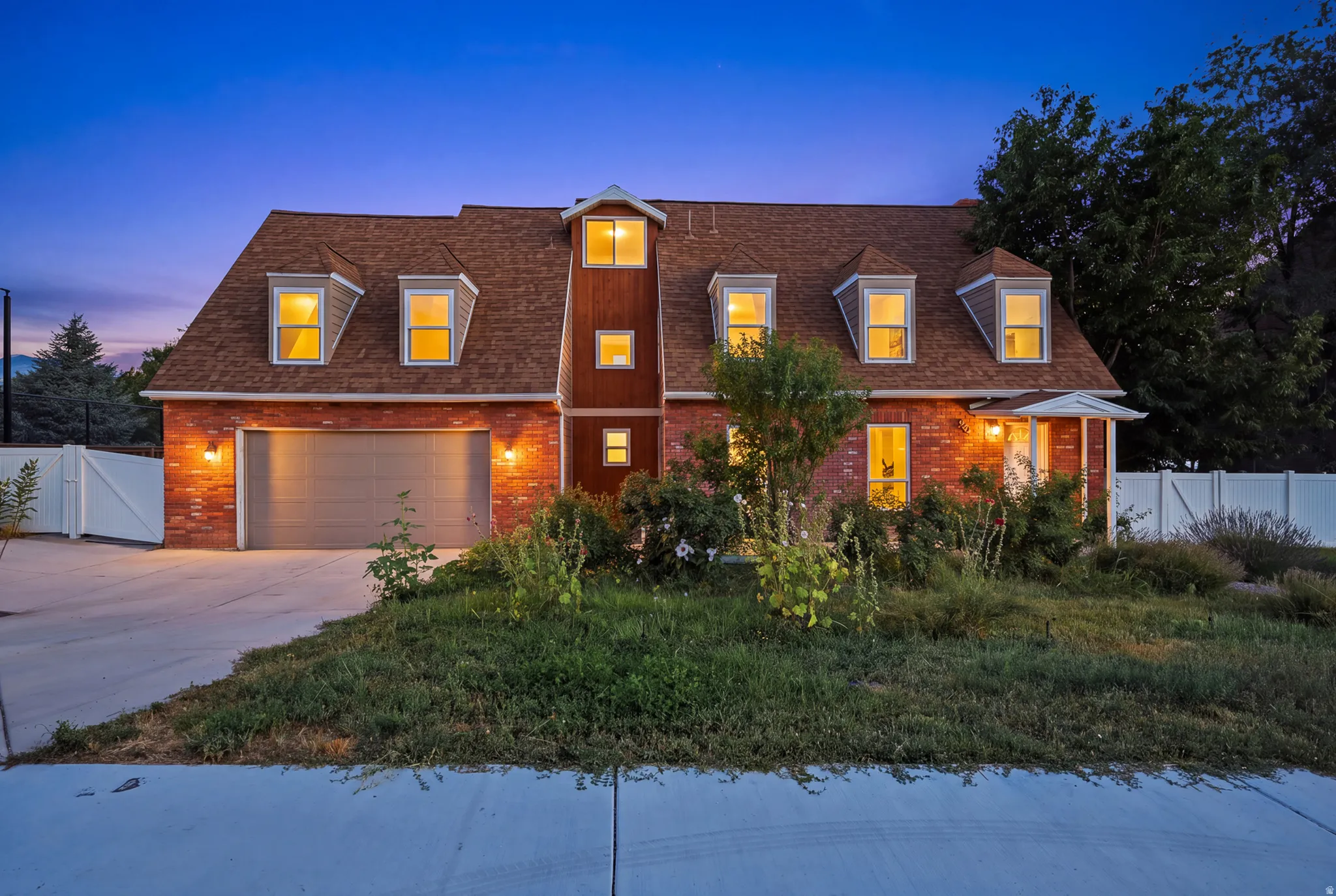 Brick exterior with dormer windows, attached garage, concrete driveway, and mature landscaping