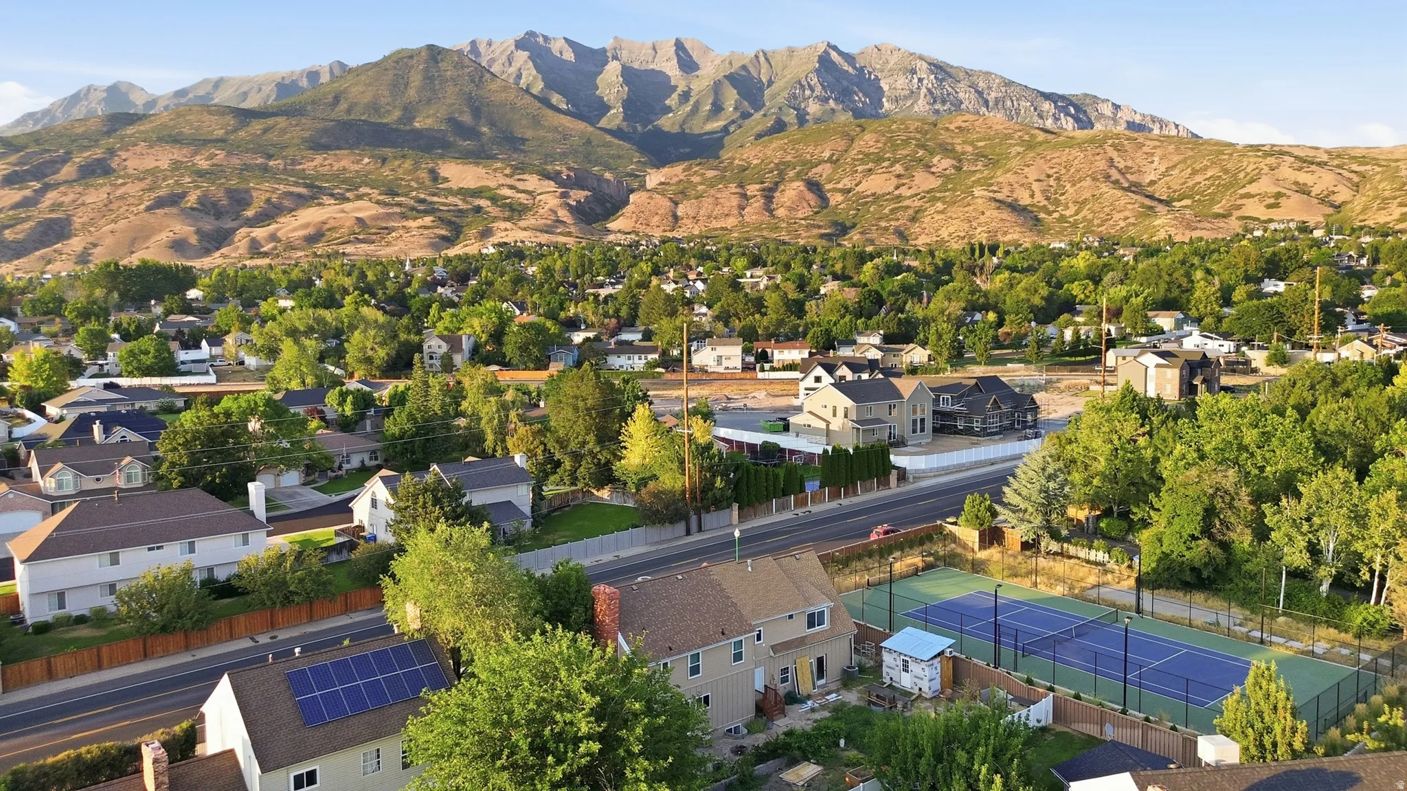 Expansive mountain range backdrop, mature trees, residential neighborhood setting, multi-lane roadway, and a tennis court with fencing