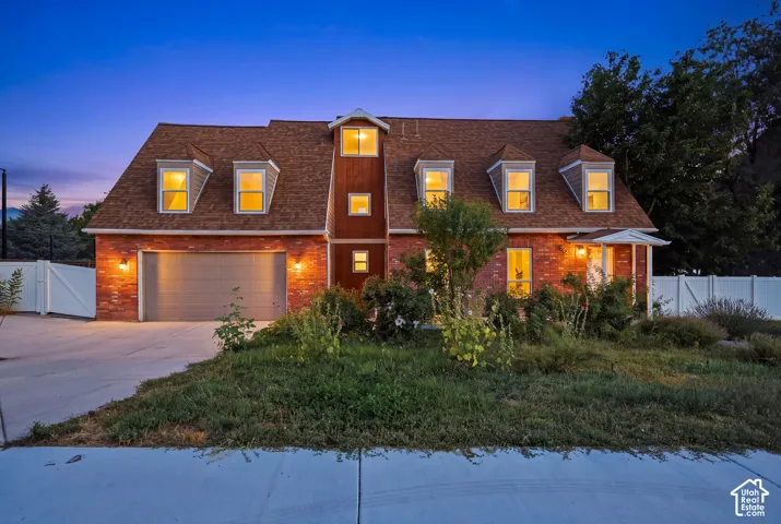 Brick exterior with dormer windows, attached garage, concrete driveway, and mature landscaping