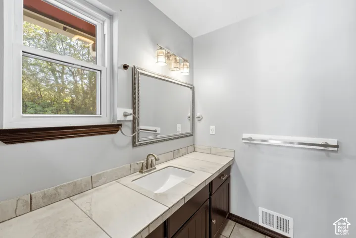 Single-pane window with wood-finish trim, tile countertop with an integrated sink, dark wood-finish vanity, and a framed mirror with overhead lighting