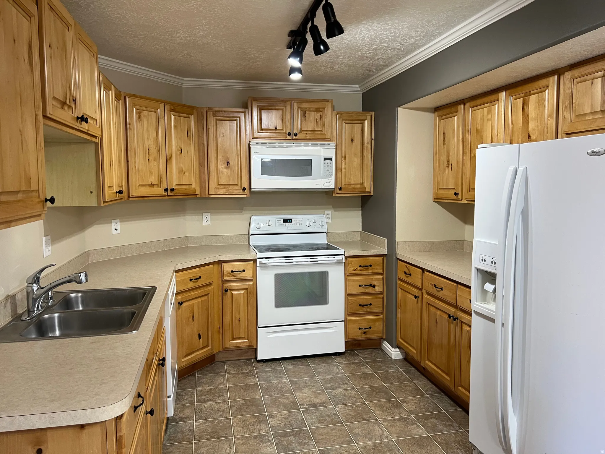 Kitchen featuring wood cabinetry, laminate countertops, a double-basin stainless steel sink, tile flooring, and white appliances