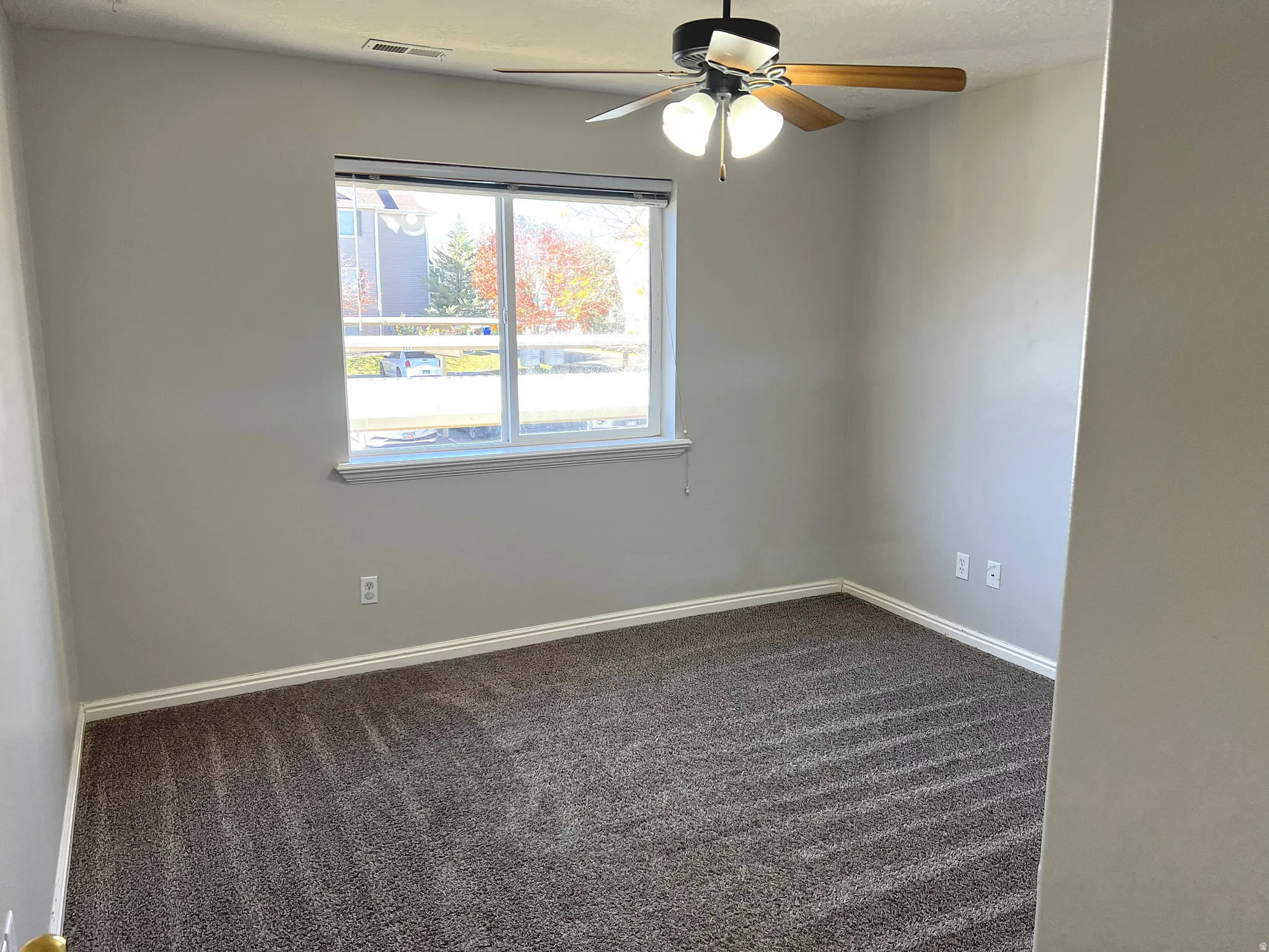 Light gray painted walls, brown patterned carpet, a single window, and a ceiling fan with integrated lighting