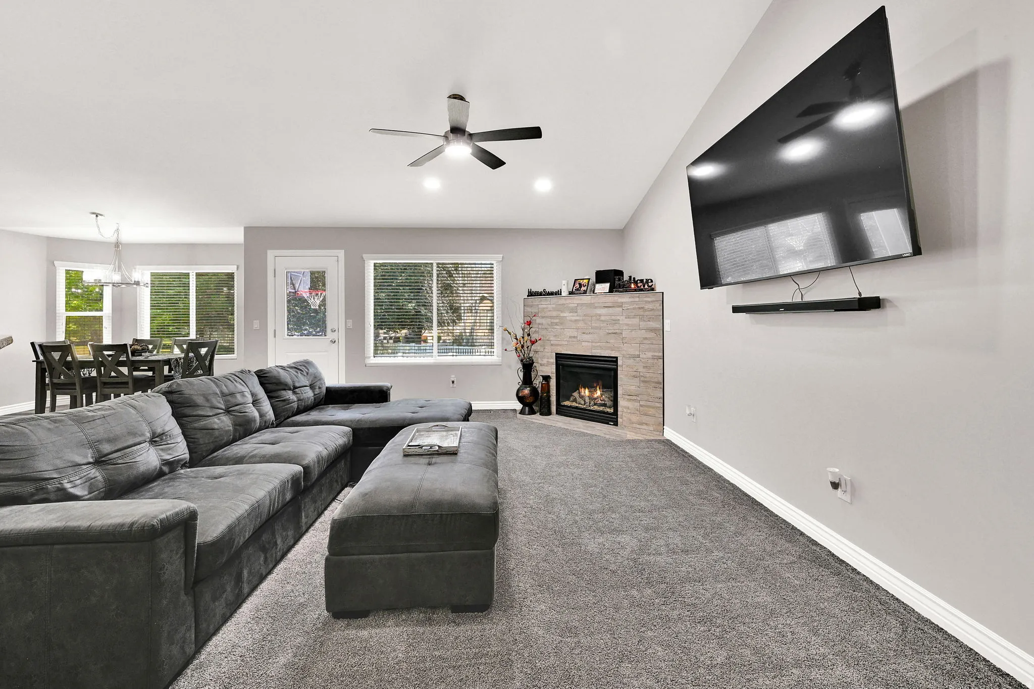 Vaulted ceiling with a ceiling fan, gas fireplace with a stone surround, large windows, and gray carpeting