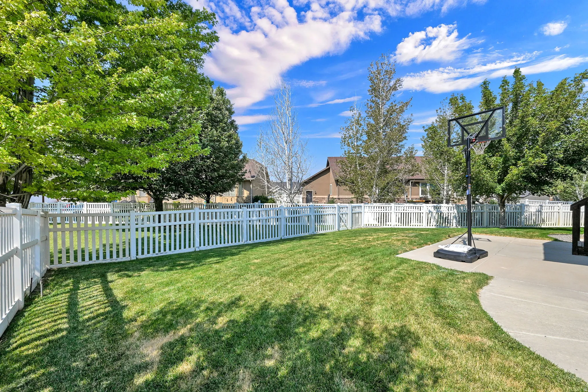 Expansive backyard featuring a manicured lawn, a white picket fence, mature trees, and a concrete patio
