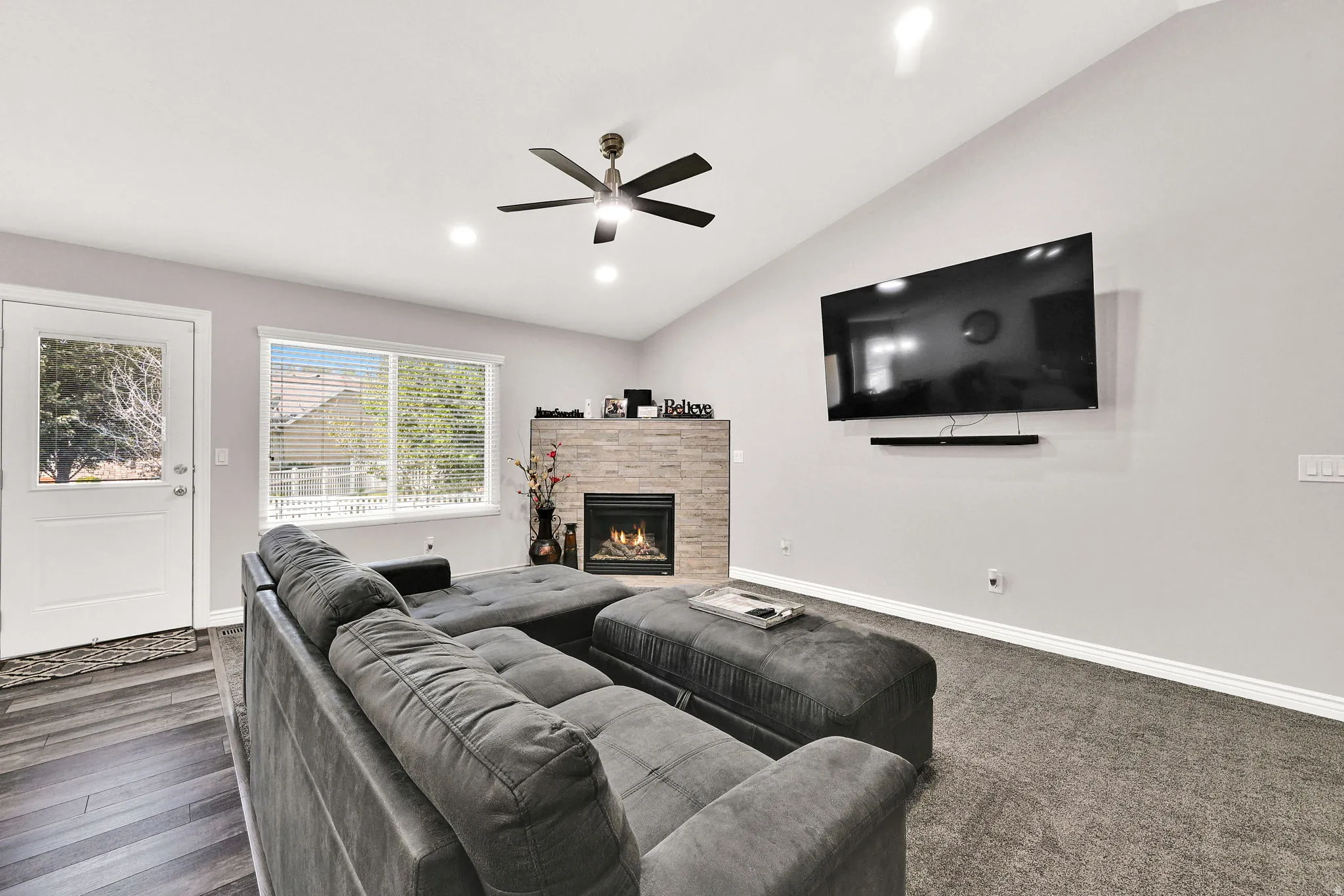 Vaulted ceiling with recessed lighting and a ceiling fan, stone-clad fireplace with a gas insert, large window with blinds, and wood-finish flooring transitioning to carpet