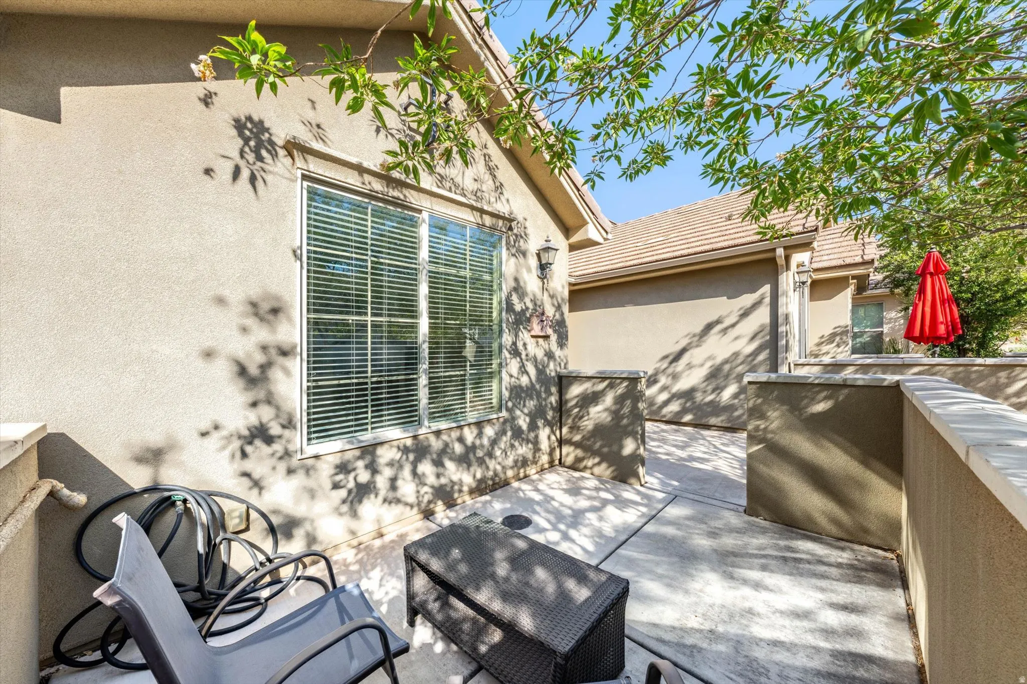 Stucco exterior with a tiled roof, featuring a concrete patio, privacy walls, and a large window with blinds