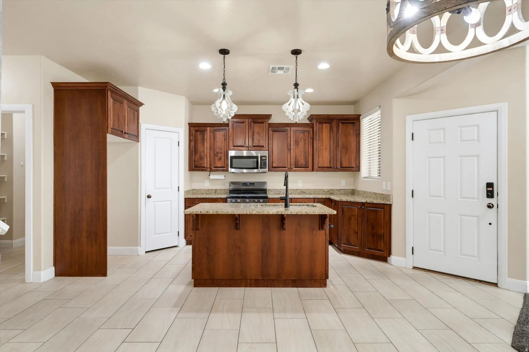 Kitchen featuring wood-finish cabinetry, a central island with a sink, granite-style countertops, stainless steel appliances, and tile flooring