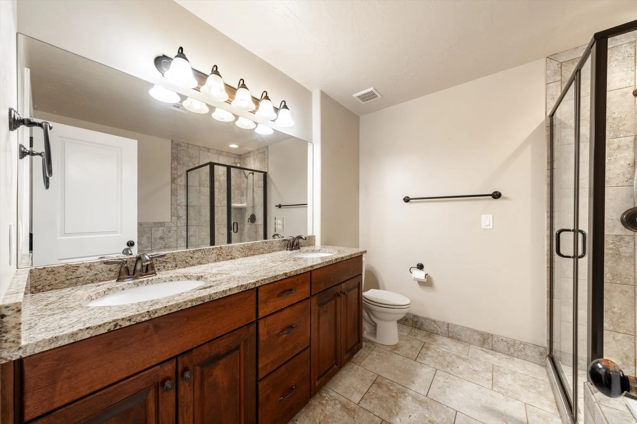 Dual vanity with stone countertop, wood-finish cabinetry, tile flooring, and a glass-enclosed shower with tile surround