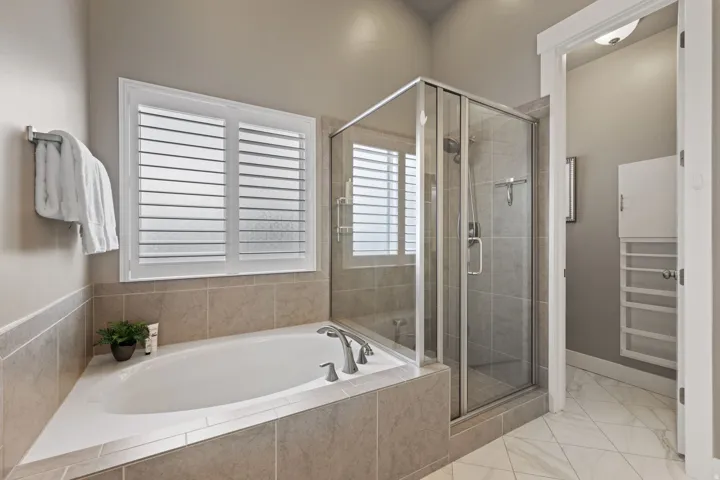 Soaking tub with tile surround, glass-enclosed shower, white plantation shutters, and large format floor tiling