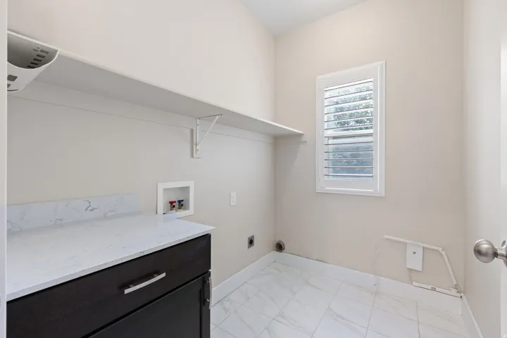 Laundry area featuring a built-in shelf, white shuttered window, dark wood-finish cabinetry, light-toned countertop, and tiled flooring