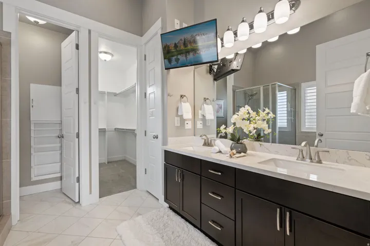 Dual vanity with undermount sinks and light-toned countertops, expansive mirror with vanity lighting, glass-enclosed shower, walk-in closet with built-in shelving, and light-toned tile flooring