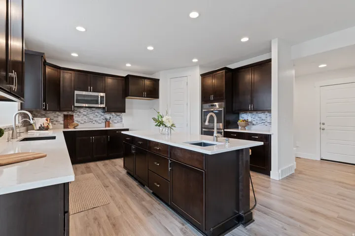 Kitchen featuring dark wood-finish cabinetry, white countertops, tile backsplash, built-in stainless steel appliances, and an island with an integrated sink