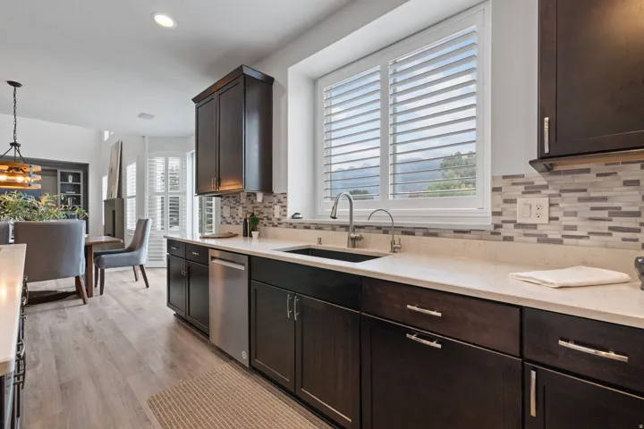 Dark wood-finish cabinetry with silver-tone hardware, light-toned countertops, stainless steel dishwasher, mosaic tile backsplash, and wood-finish flooring