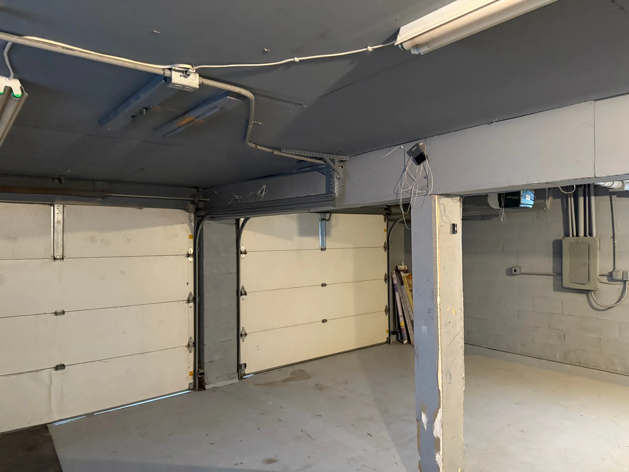 Two white segmented garage doors, exposed ceiling beams, concrete flooring, and an electrical panel