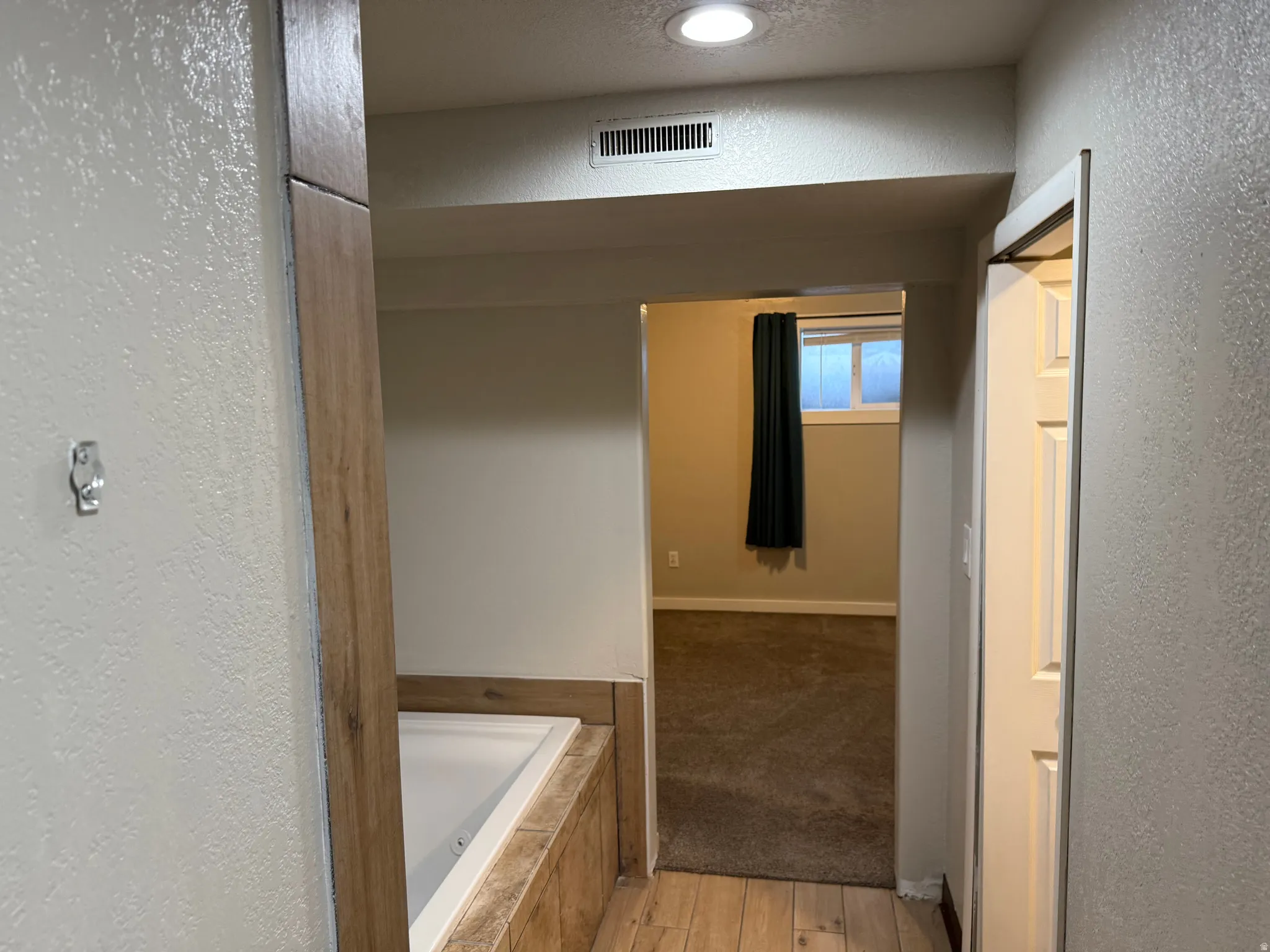 Built-in bathtub with tiled surround, wood-finish flooring, recessed lighting, and an open doorway to an adjoining carpeted room