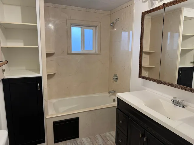 Built-in shelving, integrated bathtub with shower, dark wood-finish vanity with white countertop, framed medicine cabinet, and wood-finish flooring