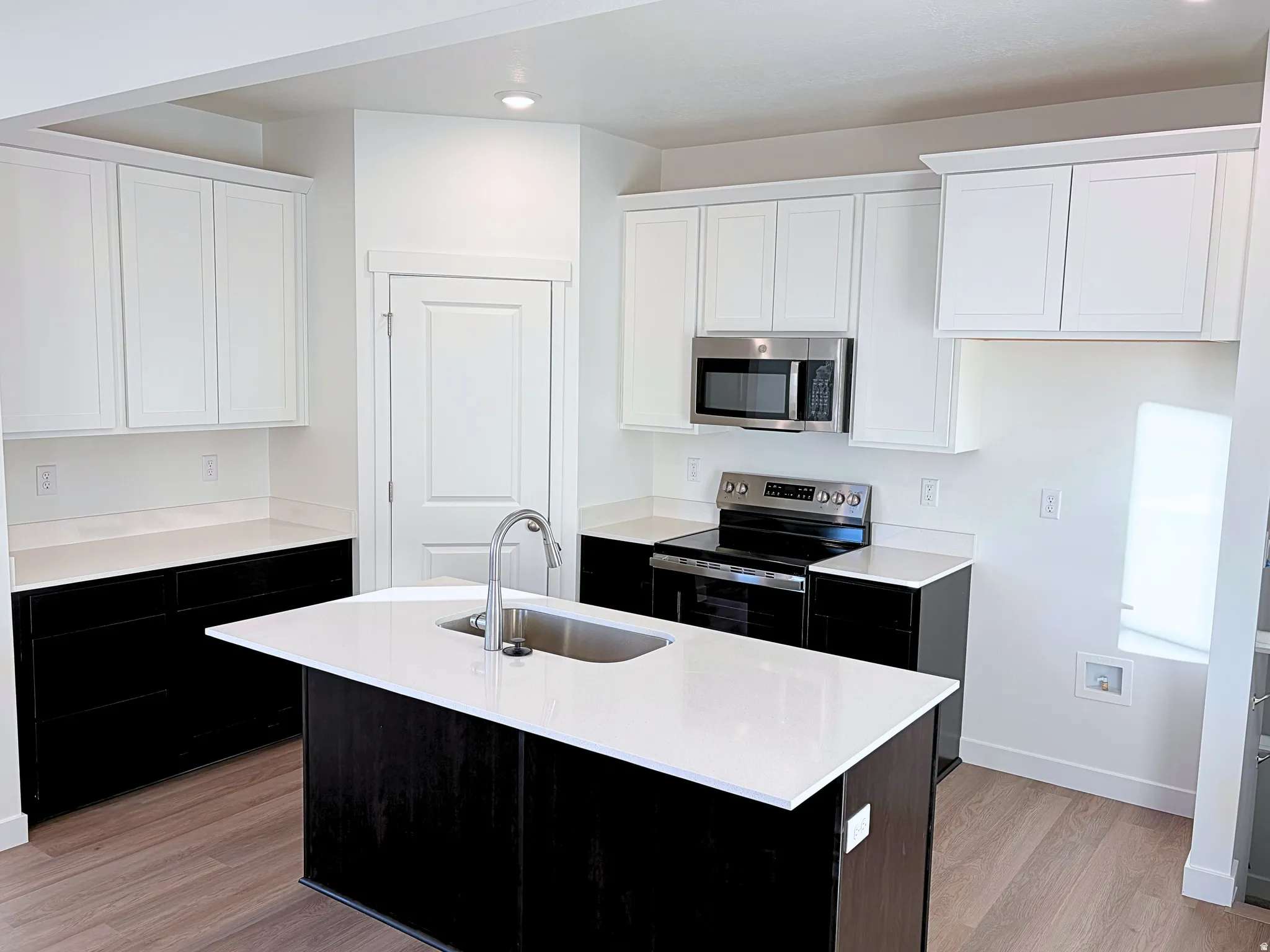 Modern kitchen featuring a central island with an undermount sink, stainless steel appliances, white upper cabinetry, and dark lower cabinetry with light countertops, complemented by wood-finish flooring