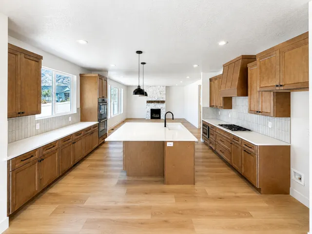 Open-concept kitchen featuring wood-finish flooring, a central island with an integrated sink, dual wall ovens, and custom cabinetry with coordinating range hood