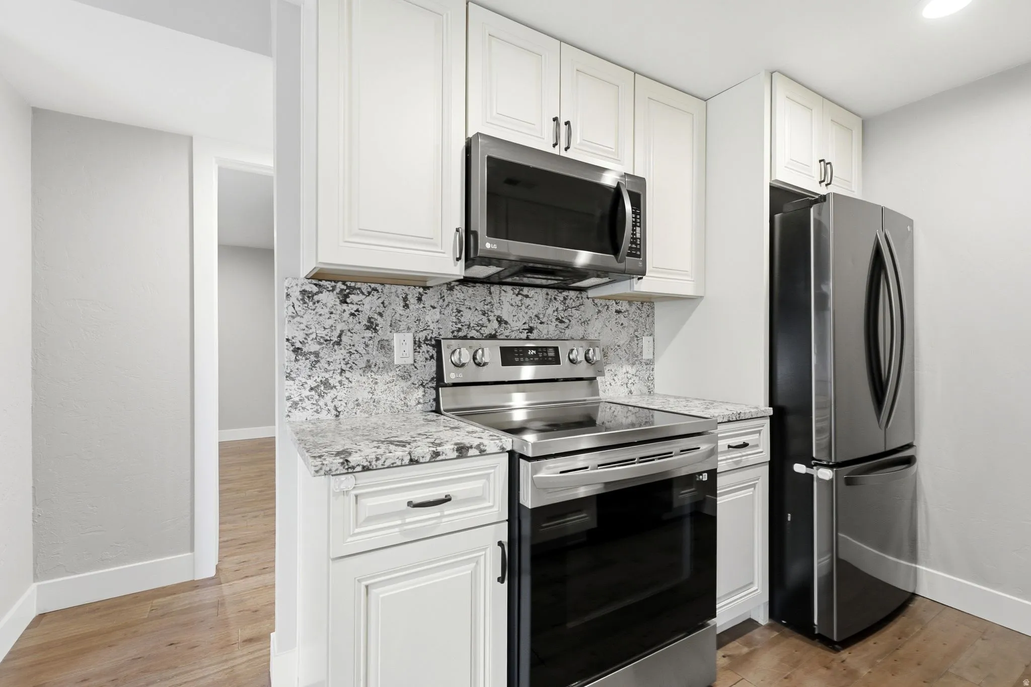 White cabinetry, stainless steel appliances, patterned stone countertops, complementing stone backsplash, and wood-finish flooring