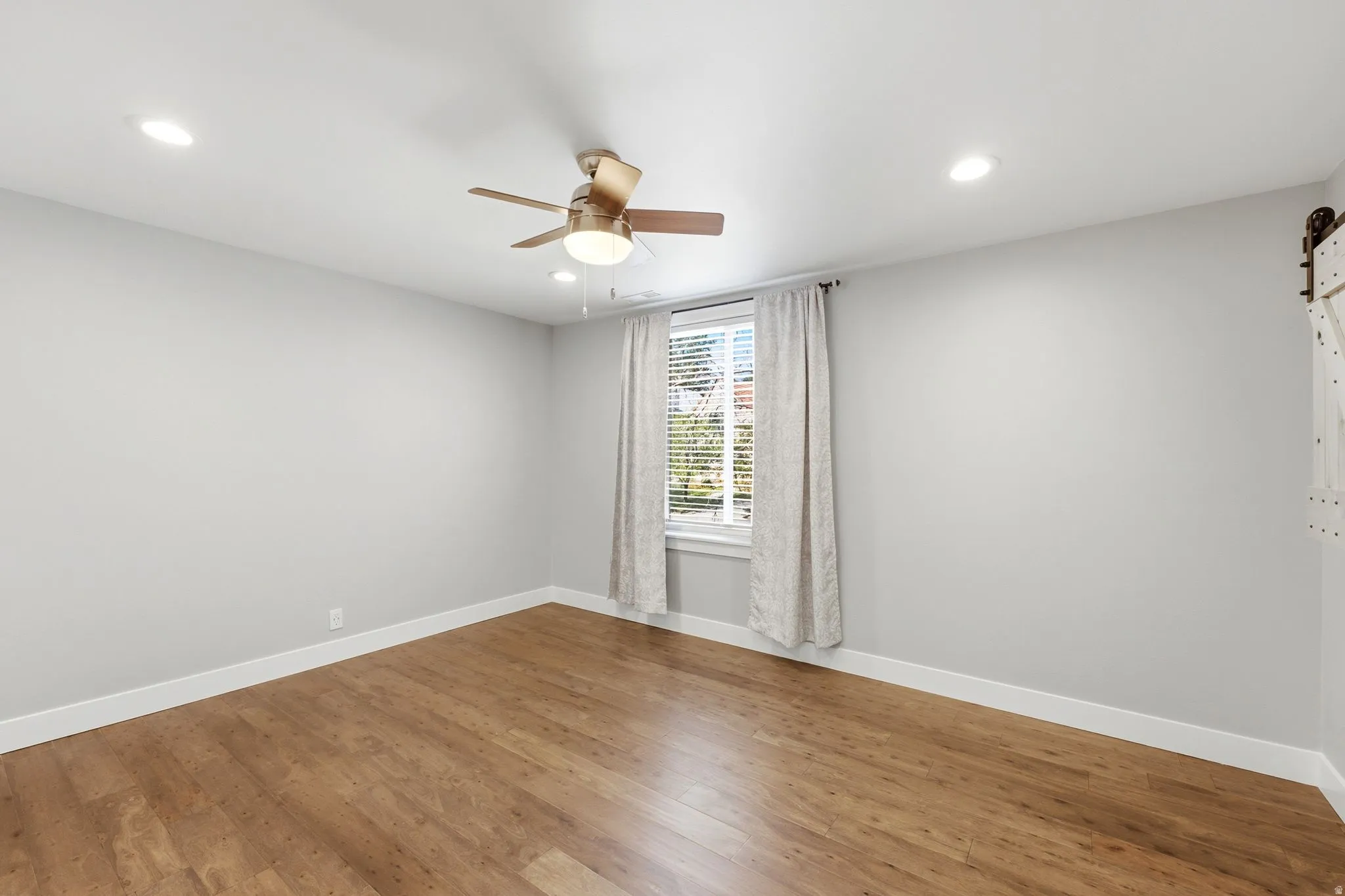 Wood-finish flooring, recessed lighting, ceiling fan, window with blinds, and a barn door hardware system