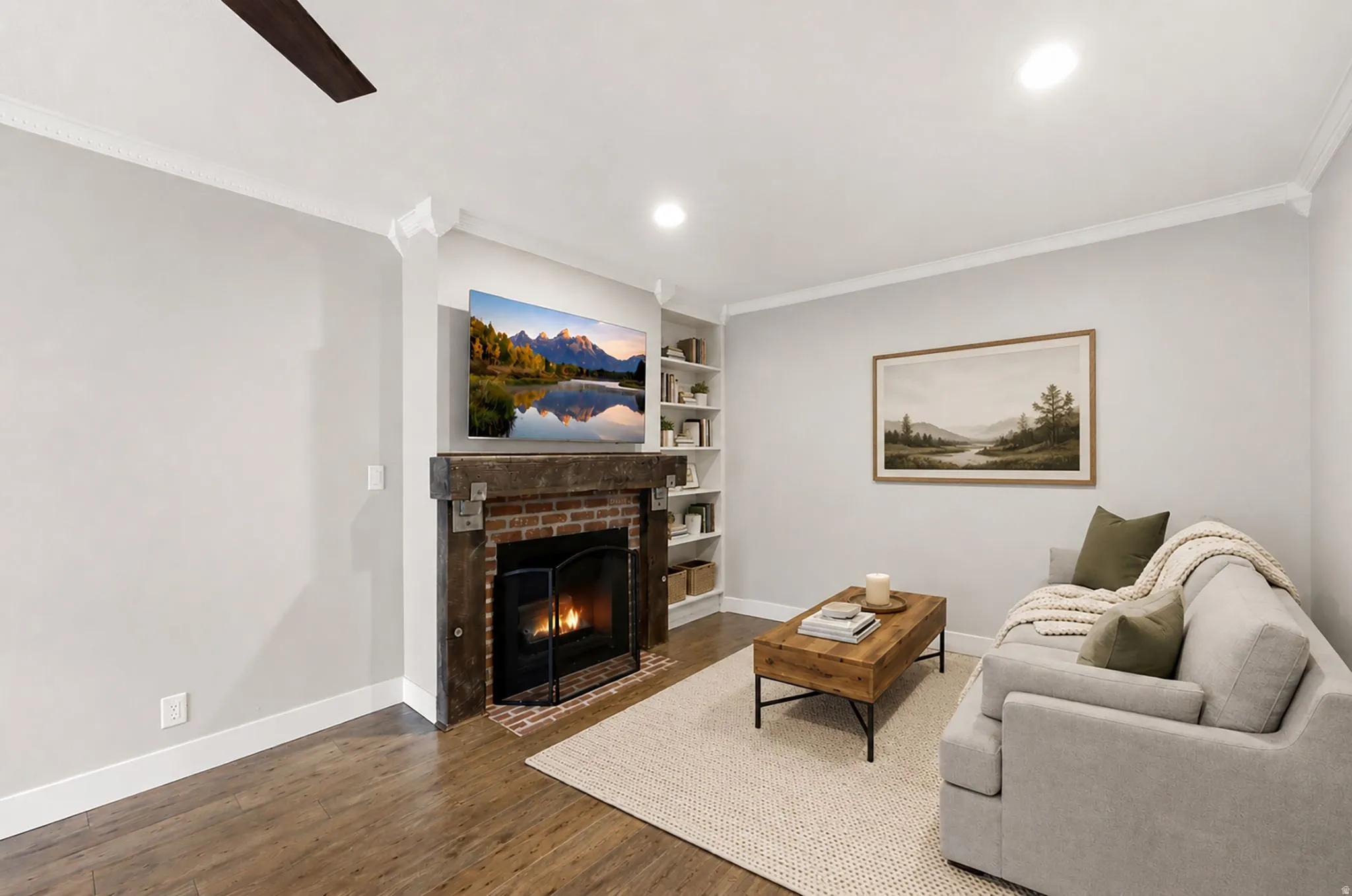 Brick-surround fireplace with a wood mantle, built-in shelving, wood-finish flooring, recessed lighting, and crown molding