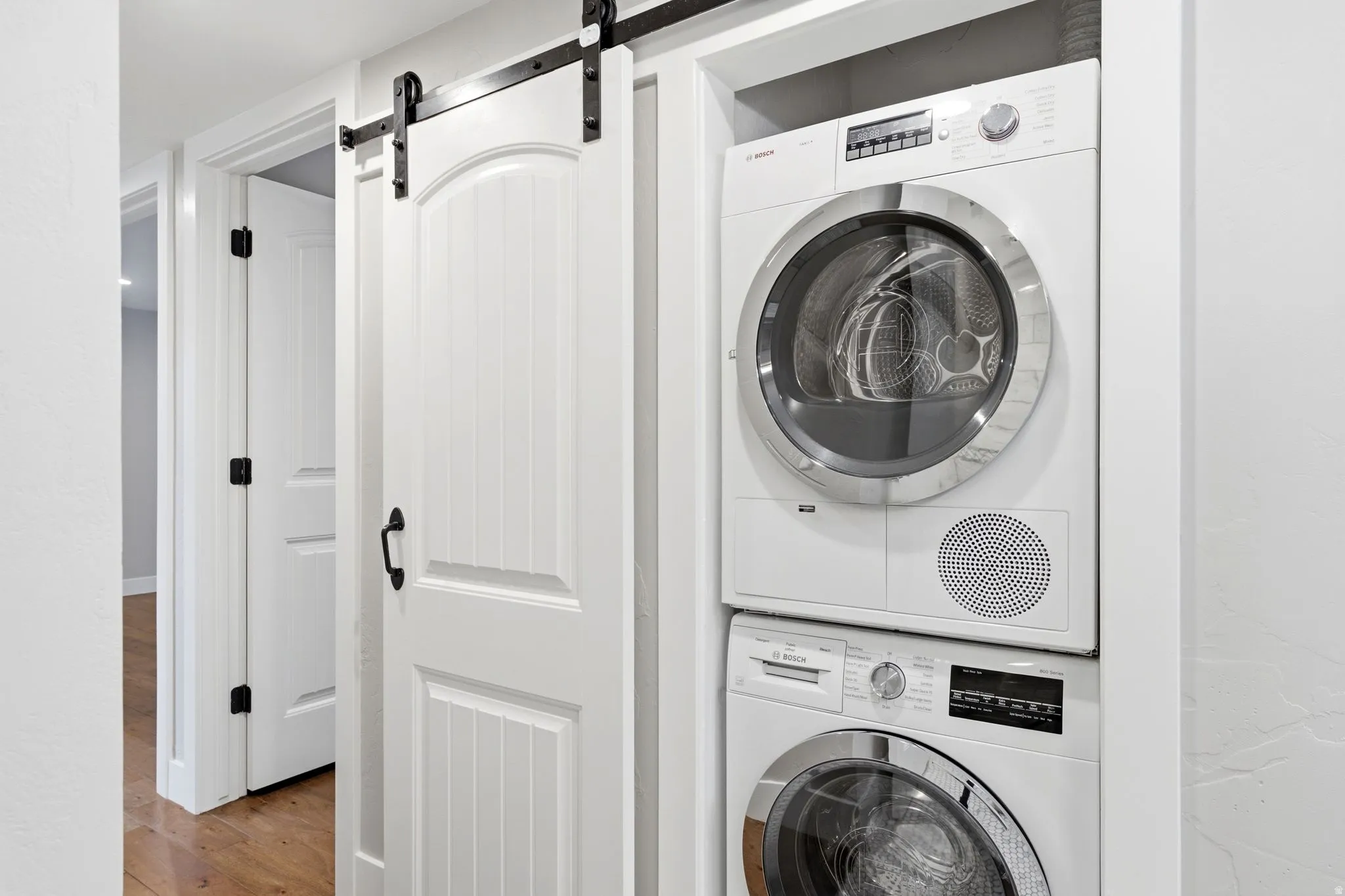 Stacked washer and dryer unit, white paneled barn door, dark hardware, and wood-finish flooring