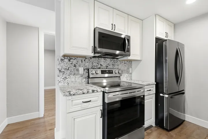 White cabinetry, stainless steel appliances, patterned stone countertops, complementing stone backsplash, and wood-finish flooring