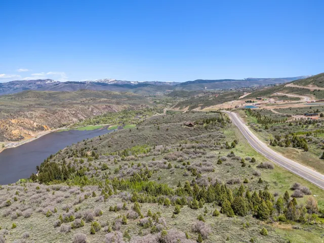 Expansive natural landscape featuring a body of water, distant snow-capped mountains, a winding paved road, and scattered evergreen trees