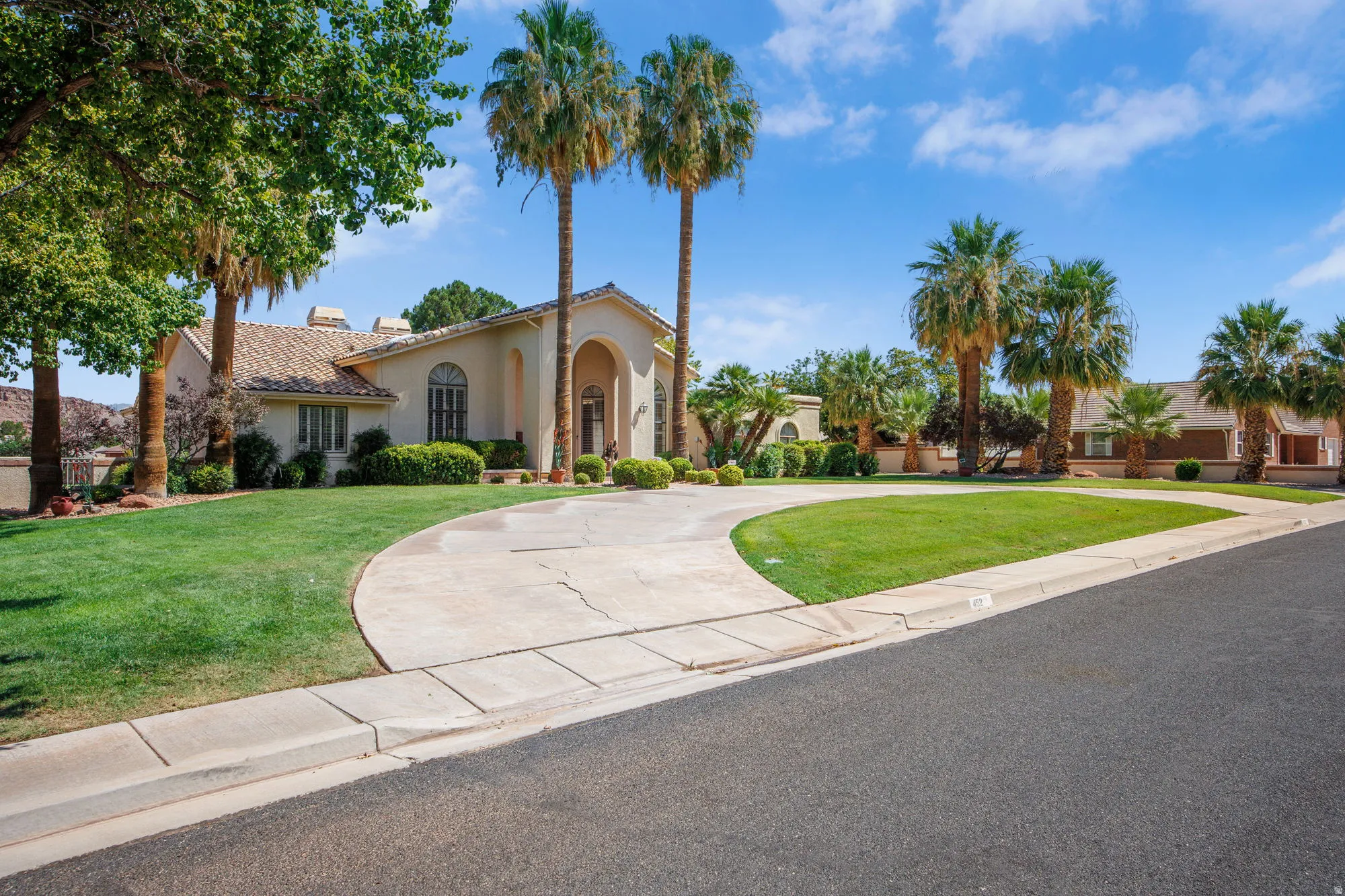 Mediterranean-inspired residence featuring a curved driveway approach, stucco exterior with arched entryway, mature palm trees and established landscaping, manicured lawn, and mature shade trees throughout the grounds