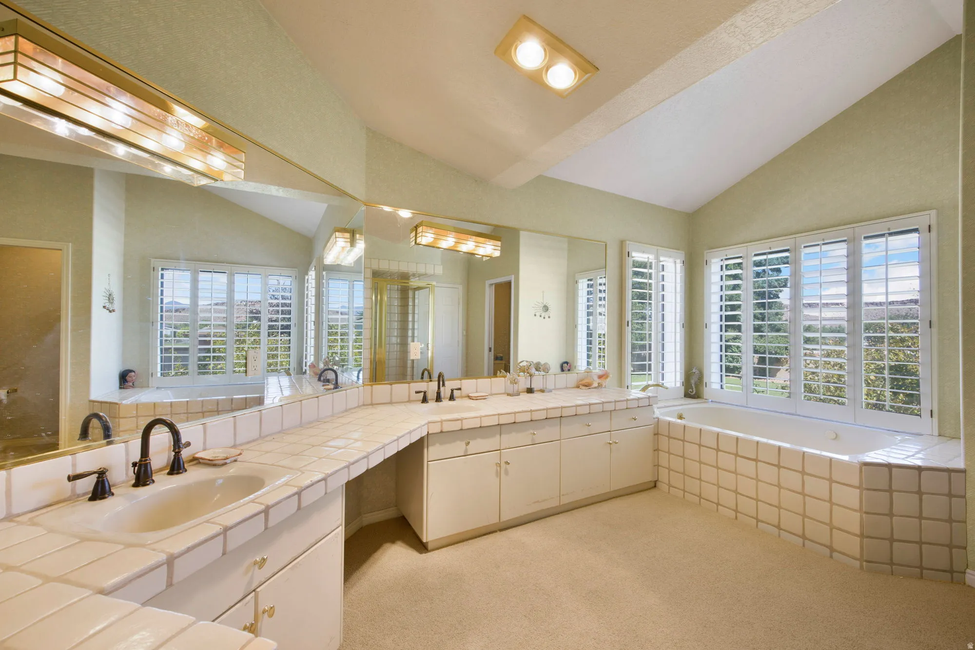 Spa-inspired primary bathroom featuring dual vanities with cream cabinetry and light-finish countertops, extensive tile work including subway tile accents, recessed ceiling lighting, and an oversized soaking tub positioned beneath expansive multi-pane win