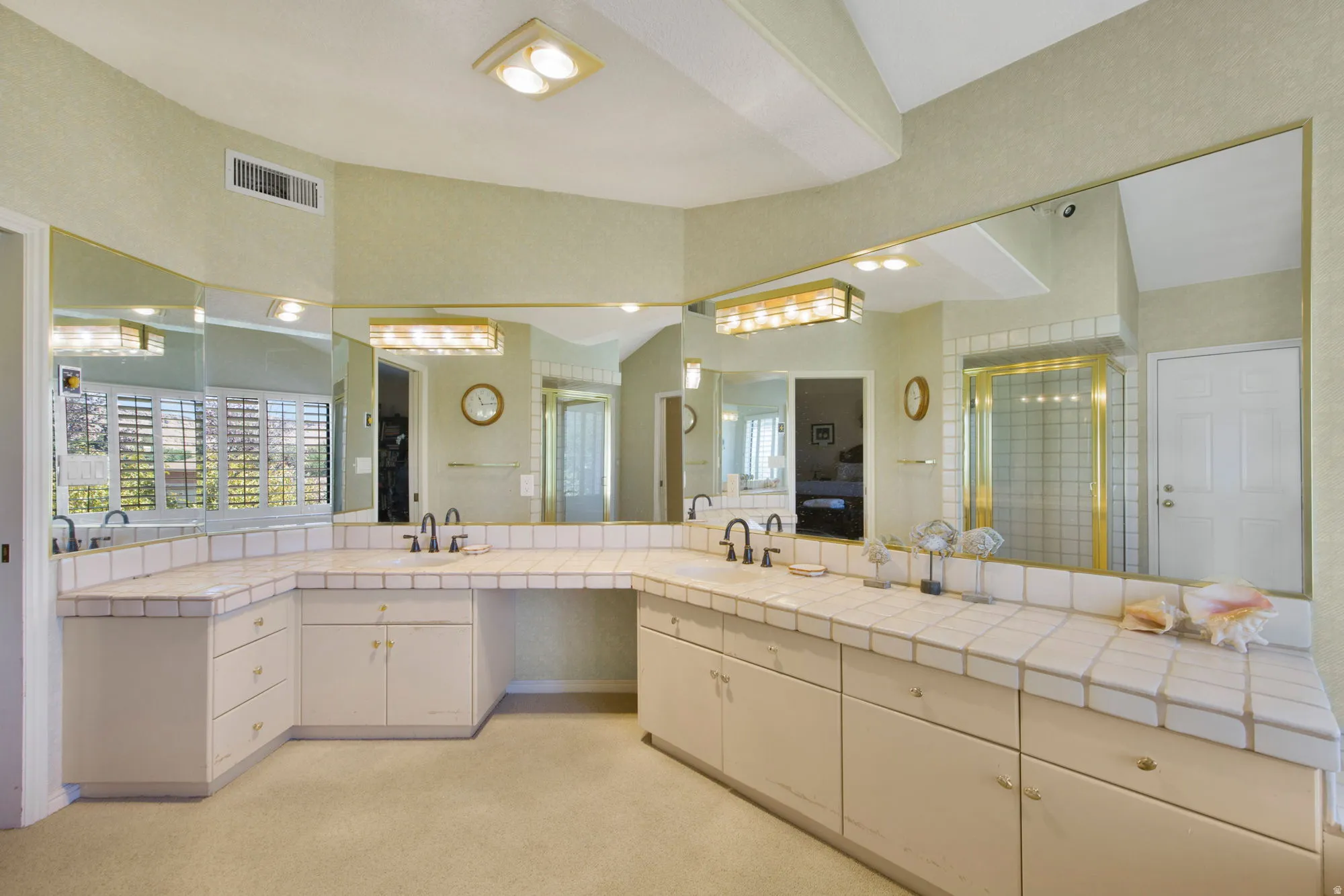 Luxurious primary bathroom suite featuring dual vanities with ample storage, extensive mirror wall with integrated lighting, tile countertops, and abundant natural light from corner window placement