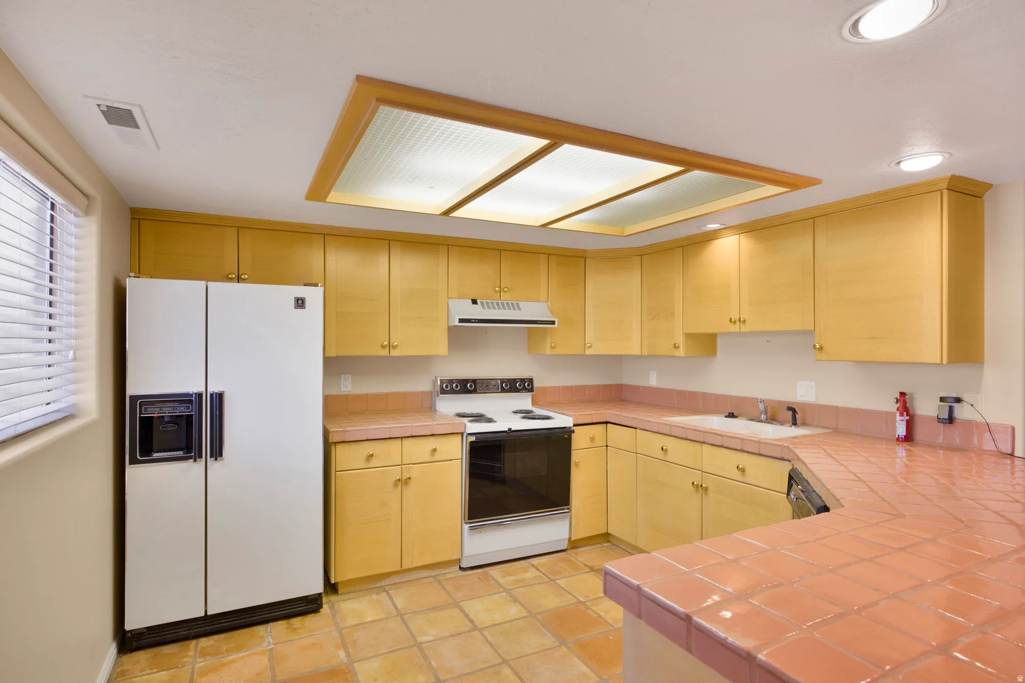 Spacious kitchen featuring honey-toned wood cabinetry, white side-by-side refrigerator, electric range with stainless steel finish, and terracotta tile flooring